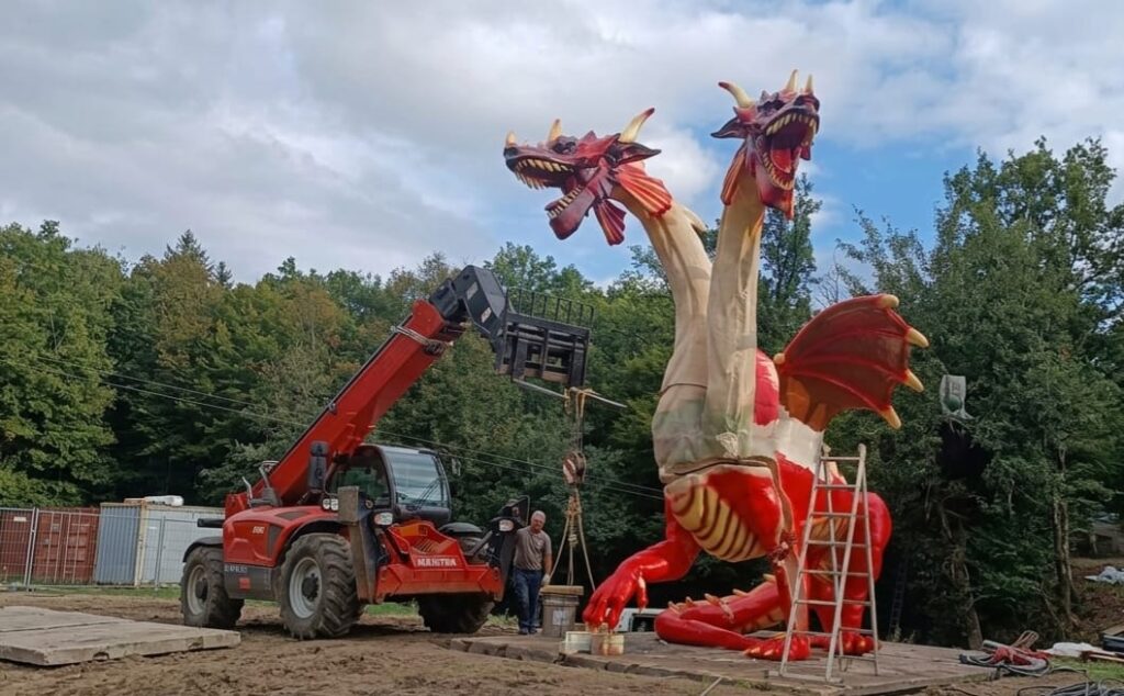Montaż trójgłowego smoka XXL przy użyciu ładowarki teleskopowej Manitou na terenie Skalka Family Park. Proces precyzyjnego pozycjonowania i kotwienia rzeźby z laminatu na betonowym fundamencie.