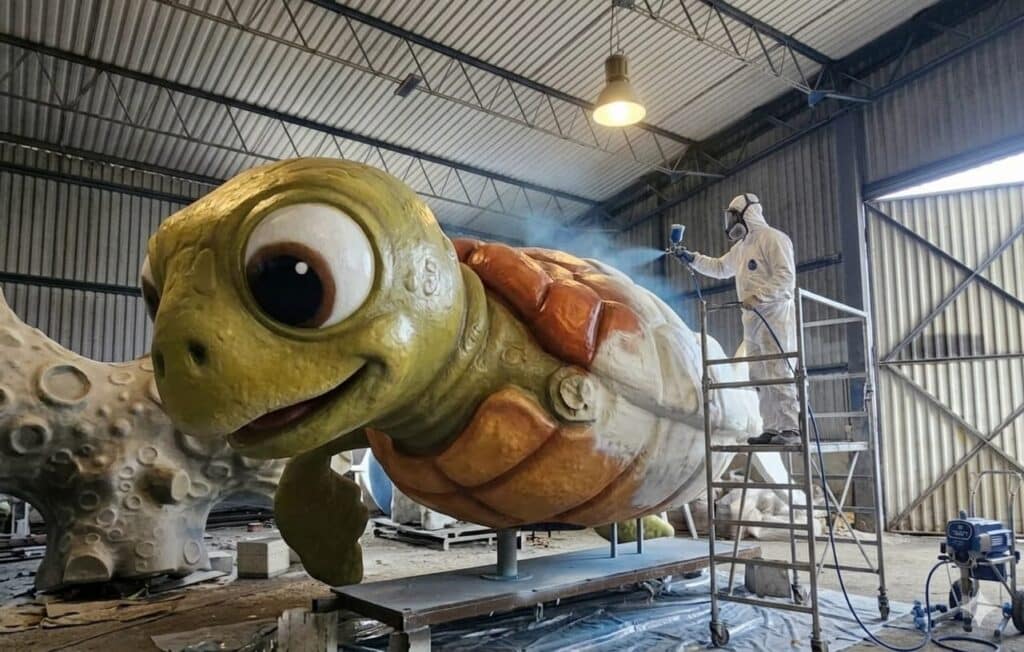 Pracownik w kombinezonie ochronnym malujący natryskowo wielkoformatową rzeźbę żółwia z laminatu w hali produkcyjnej Malpol.