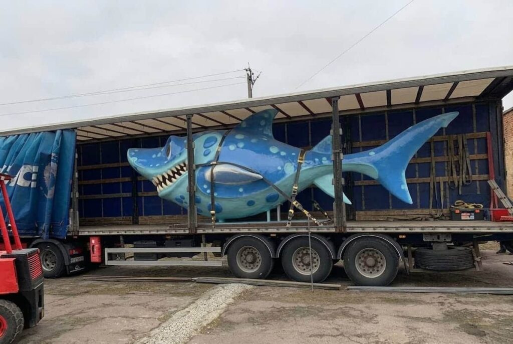 Gigantyczna niebieska rzeźba rekina na naczepie ciężarówki podczas transportu na plac budowy - profesjonalna logistyka rzeźb wielkoformatowych Malpol.