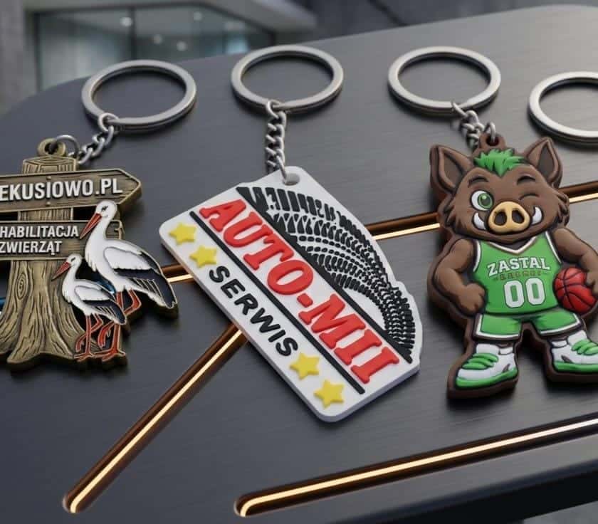 Funkcjonalne, trójwymiarowe breloki reklamowe dla marek takich jak Zastal, Klekusiowo i Auto-Mit, ułożone na ciemnym podłożu.